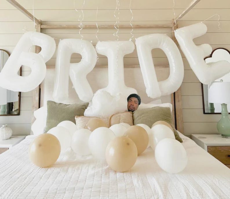 May include: A bedroom decorated for a bridal celebration. White balloons spelling "BRIDE" hang above a bed covered with white linens. Additional white and beige balloons are scattered on the bed.