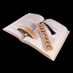 May include: A wooden bookmark and a book holder rest on an open book. The bookmark is rectangular with crescent moon cutouts and a black tassel. The book holder is curved with a circular opening and moon designs. The book is open to a page with text.