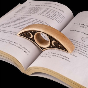 May include: A wooden ring with a crescent moon design and stars carved into the surface. The ring is resting on a book with the text "Full Moon Meditations" printed on the page.