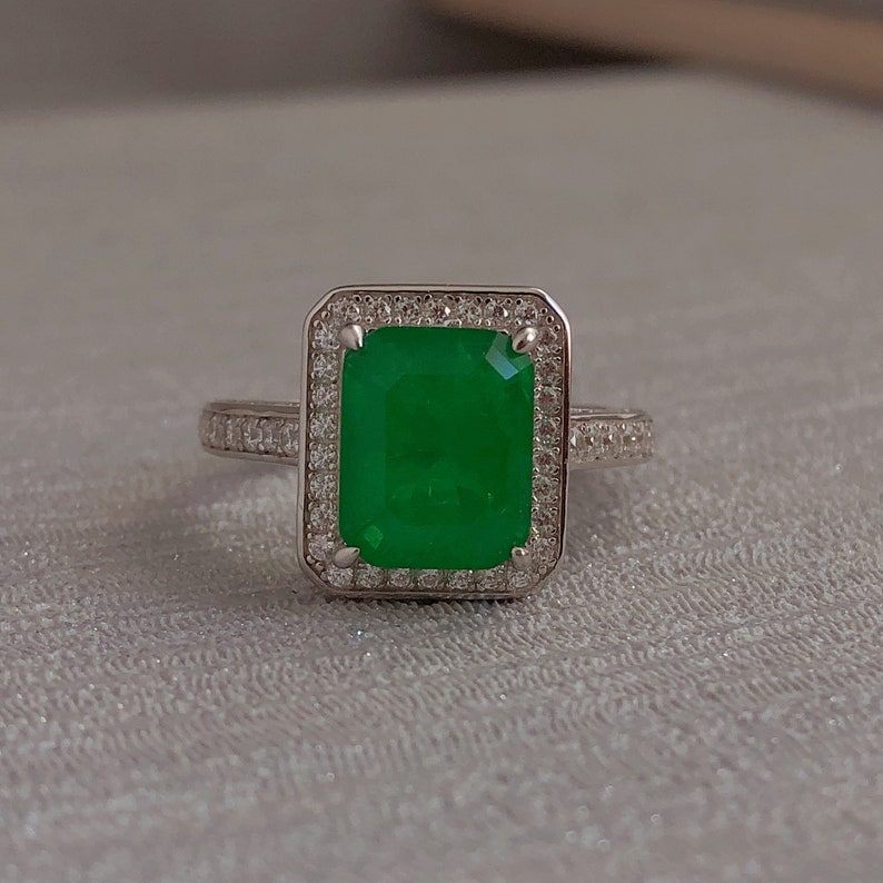 May include: A silver ring with a large emerald green gemstone surrounded by smaller clear gemstones.