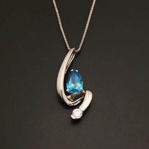 May include: A silver pendant necklace featuring a pear-shaped, bright blue gemstone. The gemstone is set within a swirling, polished silver design, with a small, round, clear stone accent. The necklace hangs on a delicate silver chain.