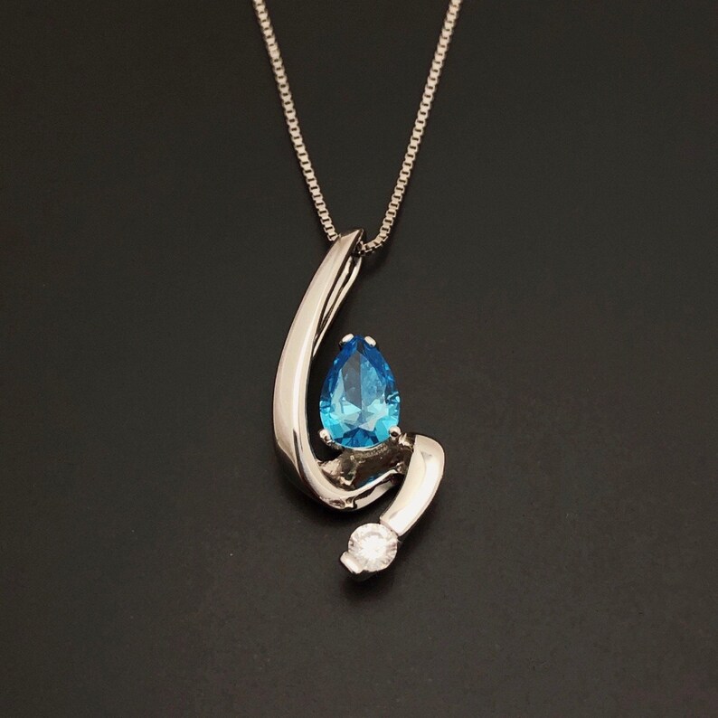May include: A silver pendant necklace featuring a pear-shaped, bright blue gemstone. The gemstone is set within a swirling, polished silver design, with a small, round, clear stone accent. The necklace hangs on a delicate silver chain.