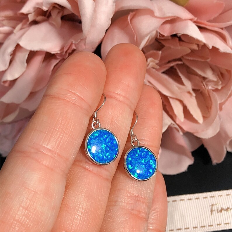 May include: A pair of round, blue opal earrings with a silver-colored border. The earrings are held up by a silver hook. The blue opal has a sparkling, glittery appearance. The background includes a pink flower and a black surface.