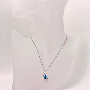 May include: A silver necklace with a teardrop-shaped pendant. The pendant features a large, bright blue gemstone and a smaller, clear gemstone. The necklace is displayed on a white mannequin.
