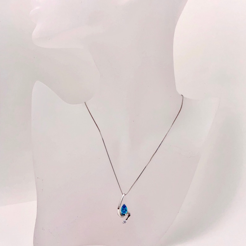May include: A silver necklace with a teardrop-shaped pendant. The pendant features a large, bright blue gemstone and a smaller, clear gemstone. The necklace is displayed on a white mannequin.