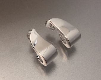 May include: A pair of polished silver stud earrings. Each earring has a curved, ribbon-like design with a smooth, reflective surface. The earrings are set against a light gray background, highlighting their minimalist and modern aesthetic.