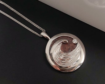 Puede incluir: Collar de plata con un colgante redondo que presenta un diseño de ola y una pequeña piedra transparente. El colgante está sobre un fondo marrón.