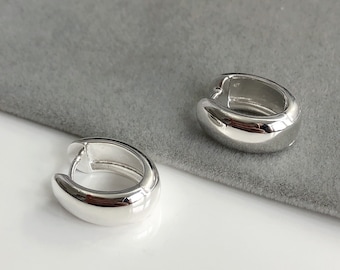 May include: Two silver-colored, chunky hoop earrings. The earrings have a smooth, rounded design and a secure clasp closure. The earrings are displayed on a gray surface, reflecting light.