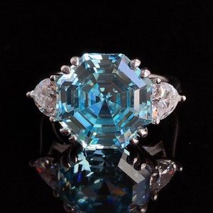 May include: A silver ring with a large, light blue, octagonal-cut central gemstone, accompanied by two pear-shaped, clear gemstones. The ring is displayed against a black backdrop, accentuating the brilliance of the stones.
