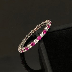 Peut inclure: Une bague en argent ornée de pierres précieuses roses et claires alternées. La bague est présentée sur un fond noir, mettant en valeur l'éclat des pierres. Les pierres précieuses sont rondes et espacées uniformément autour de l'anneau.