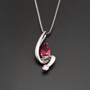 Könnte beinhalten: Eine silberne Halskette mit einem birnenförmigen roten Edelstein und einem kleineren, klaren Edelstein. Der Anhänger hängt an einer zarten silbernen Kette.