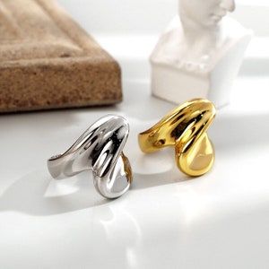 May include: Two abstract rings, one gold and one silver, with a wavy, organic design. The rings are on a white surface.