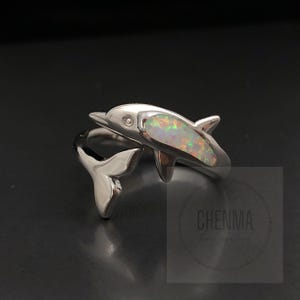 May include: A silver dolphin ring featuring an opal inlay. The dolphin's body is a bright, iridescent opal, and the ring wraps around the finger with the dolphin's tail. The ring is set against a dark background. The text "CHENMA Fine Jewellery" is visible.