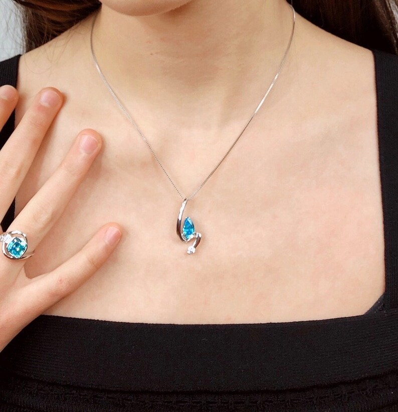 May include: A silver necklace with a pendant featuring a teardrop-shaped, light blue gemstone and a small, round, clear gemstone. A matching ring with a similar design is also visible.