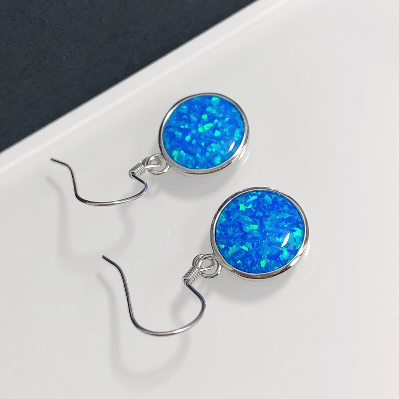 May include: A pair of round, blue opal earrings with a silver-colored metal setting. The earrings have a hook closure. The opal stones have a vibrant blue color with flecks of green.