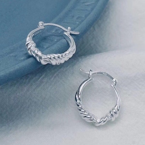 May include: A pair of silver hoop earrings. Each earring features a twisted rope design along the lower half of the hoop. The earrings are displayed on a blue and white surface, with a soft, blurred background.