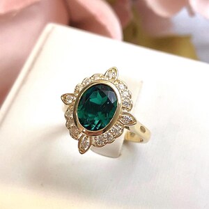 May include: A gold ring featuring a large, oval-cut emerald-green gemstone. The gemstone is surrounded by a halo of small, clear gemstones and decorative gold accents. The ring has a classic, elegant design.