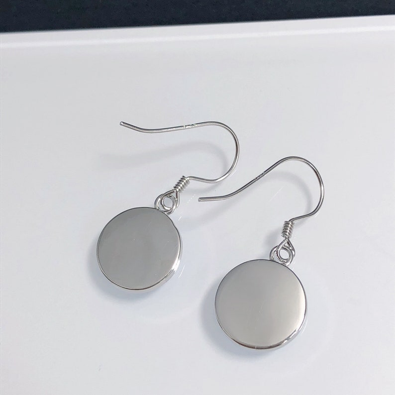 May include: A pair of silver-colored dangle earrings. Each earring features a circular, polished disc suspended from a curved ear wire. The ear wires have a coiled design near the disc. The earrings are displayed on a white surface.
