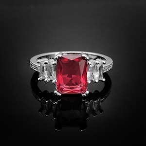 Op de afbeelding: Een zilveren ring met een grote, rode, smaragd geslepen edelsteen omringd door kleinere, heldere, rechthoekige edelstenen.