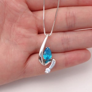 May include: A silver pendant necklace featuring a teardrop-shaped, bright blue gemstone and a small, round, clear gemstone. The pendant has a flowing, abstract design. The necklace chain is a delicate silver box chain.
