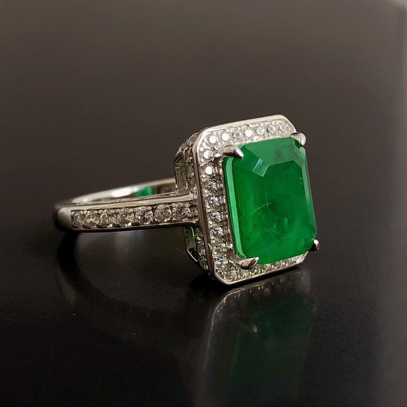 May include: A silver ring with a large emerald green gemstone surrounded by smaller clear gemstones.