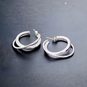 Op de afbeelding: Een paar zilverkleurige oorringen. Elke oorring heeft een drievoudig bandontwerp dat elkaar kruist. De oorbellen hebben een stekersluiting en worden op een donkere ondergrond gepresenteerd. De oorbellen zijn een modern sieradenaccessoire.