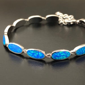 Op de afbeelding: Een zilveren armband met ovale, blauwe opaalstenen. De armband heeft een sluiting.
