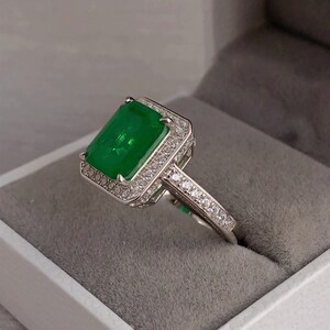 May include: A silver ring with a large emerald green gemstone surrounded by smaller clear gemstones. The ring is set in a square halo setting.