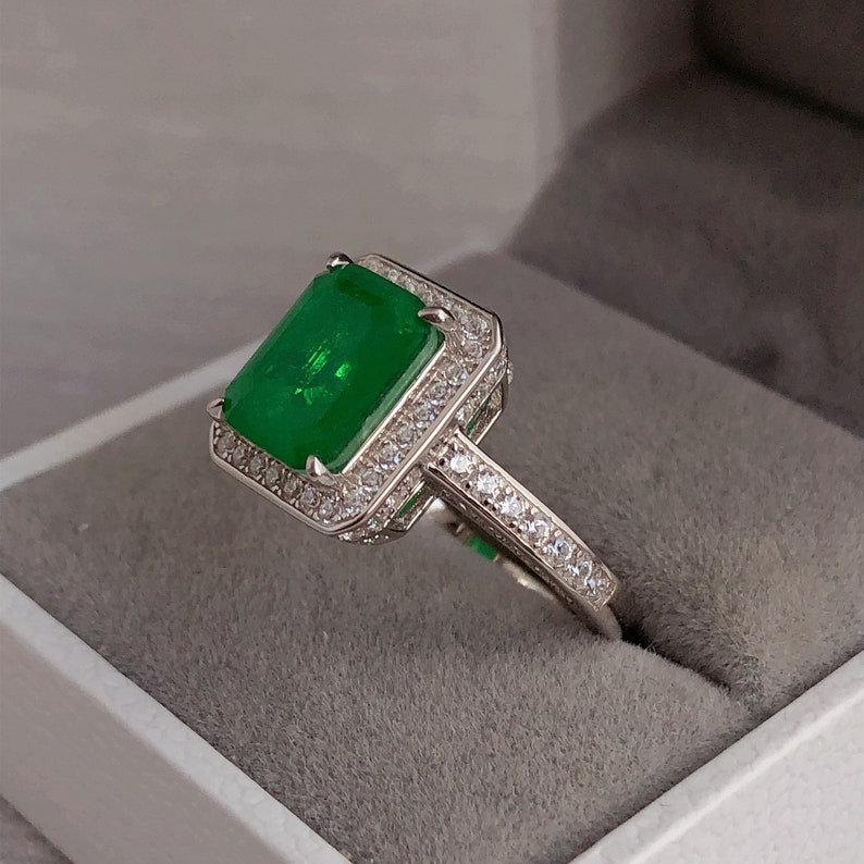 May include: A silver ring with a large emerald green gemstone surrounded by smaller clear gemstones. The ring is set in a square halo setting.