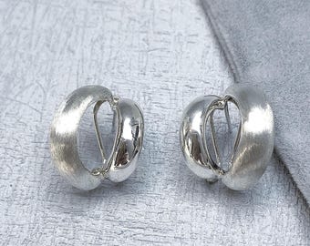 May include: A pair of silver-toned hoop earrings. Each earring has a rounded, brushed metal outer surface and a polished inner surface. The earrings are displayed on a light gray surface.