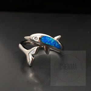 May include: A silver ring featuring a dolphin design with a blue opal stone. The dolphin's tail is curled around the ring band.
