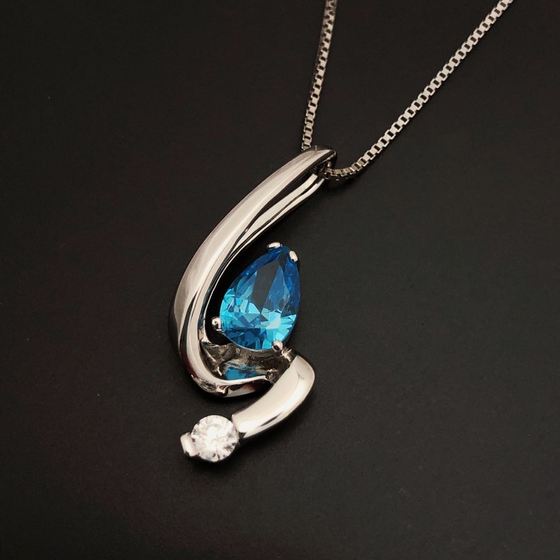 May include: A silver pendant necklace with a teardrop-shaped, bright blue gemstone. The pendant has a swirling, polished metal design and a small, round, clear stone at the bottom. The necklace chain is a delicate silver color.