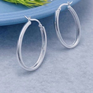May include: A pair of oval-shaped silver hoop earrings. The earrings are made of a shiny, reflective metal and have a simple, classic design. The hoops are shown against a light blue background.
