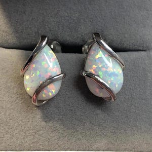 Puede incluir: Un par de pendientes de plata en forma de lágrima con piedras de ópalo blanco. Los pendientes tienen un diseño delicado y retorcido.