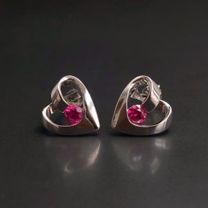 Op de afbeelding: Een paar zilveren hartvormige oorbellen met een roze edelsteen in het midden van elk hart.