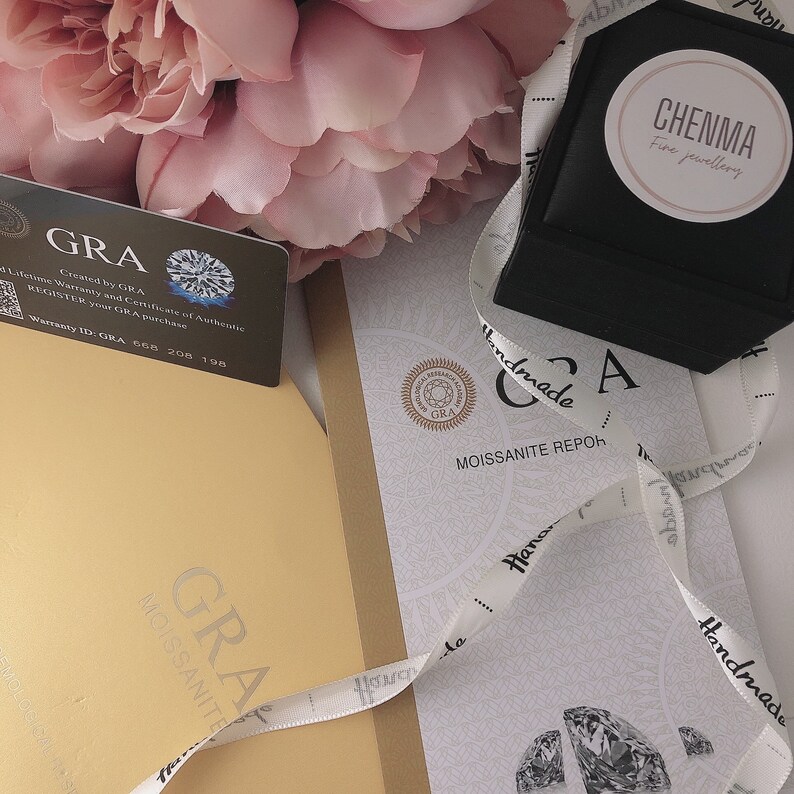 May include: A white card with the text "GRA" in black, a diamond symbol, and the text "Lifetime Warranty and Certificate of Authentic" in black. The card is on a brown surface with a white ribbon with the text "Handmade" in black. The ribbon is also on a white card with the text "GRA" in black, a diamond symbol, and the text "Moissanite Repoh" in black.