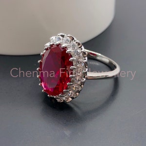 Op de afbeelding: Een zilveren ring met een grote ovale rode edelsteen omringd door kleinere heldere edelstenen.