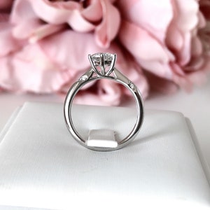May include: A silver engagement ring with a large, round, clear center stone and smaller, clear stones on the band. The ring is sitting on a white box.