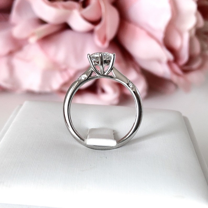 May include: A silver engagement ring with a large, round, clear center stone and smaller, clear stones on the band. The ring is sitting on a white box.