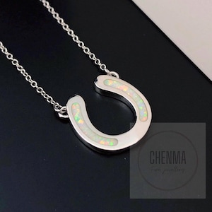 Puede incluir: Un collar con colgante de herradura de plata con incrustaciones de ópalo blanco. La herradura está suspendida de una delicada cadena. El ópalo tiene una apariencia iridiscente y colorida. El collar se muestra sobre un fondo oscuro.