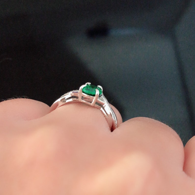 Puede incluir: Un anillo de plata con una piedra preciosa verde engastada en garras. El anillo est&aacute; en un dedo.