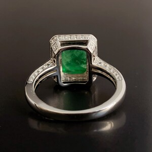 May include: A silver ring with a large emerald green gemstone set in a square halo of smaller diamonds. The ring band is also set with smaller diamonds.