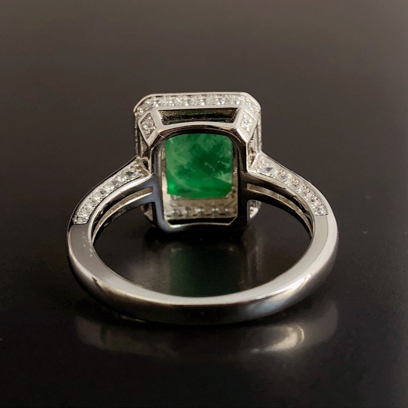 May include: A silver ring with a large emerald green gemstone set in a square halo of smaller diamonds. The ring band is also set with smaller diamonds.