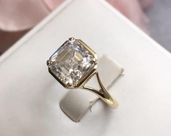 May include: A gold ring with a large, clear, square-shaped gemstone. The ring has a simple, elegant design.