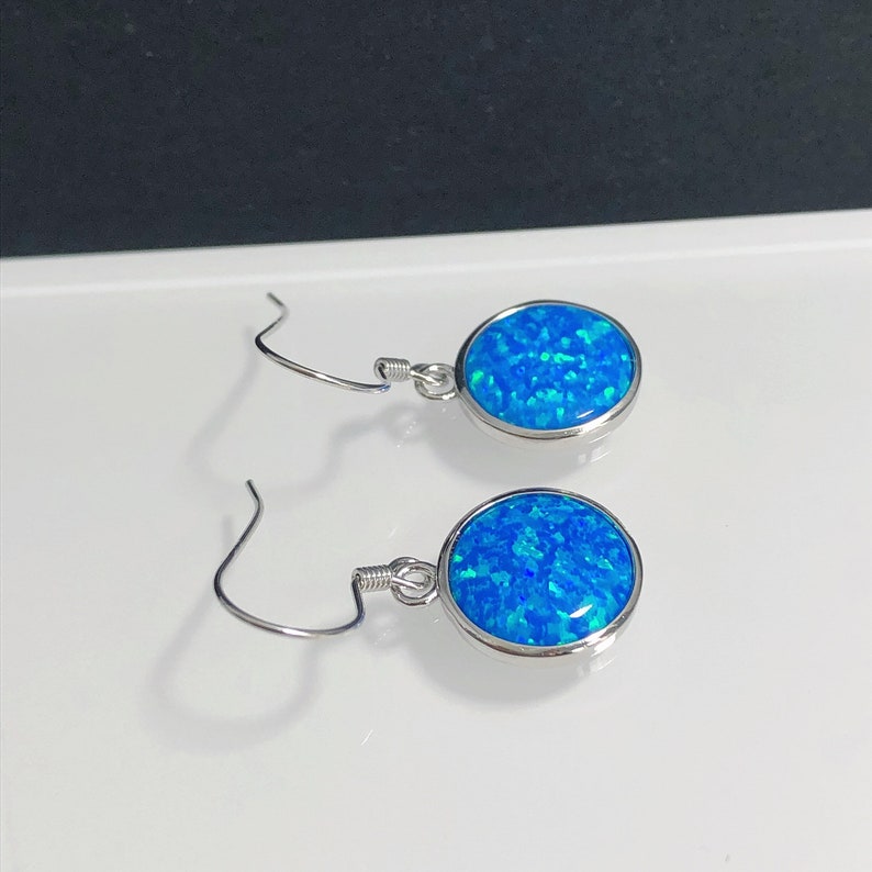 May include: A pair of silver-toned drop earrings. Each earring features a round, bright blue opal stone with a shimmering, speckled appearance. The stones are set in a silver bezel and suspended from a simple hook.