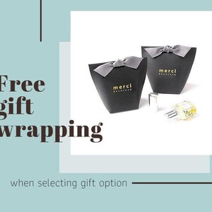 May include: Two black gift boxes with gray bows, each bearing the gold text "merci beaucoup." A perfume bottle is next to the boxes. The words "Free gift wrapping" are displayed in the image.