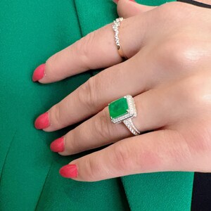 May include: A silver ring with a large emerald green gemstone surrounded by smaller diamonds. The ring has a delicate, intricate design.