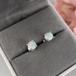 May include: A pair of silver stud earrings with white opal gemstones. The earrings are displayed in a gray velvet jewelry box.