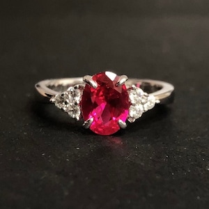 Puede incluir: Un anillo de plata con una gran piedra de rubí ovalada rodeada de piedras blancas más pequeñas.