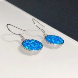 May include: A pair of silver-toned drop earrings with oval-shaped, bright blue opal stones. The opals have a shimmering, multi-tonal appearance, with hints of green within the blue. The earrings are displayed on a white surface.